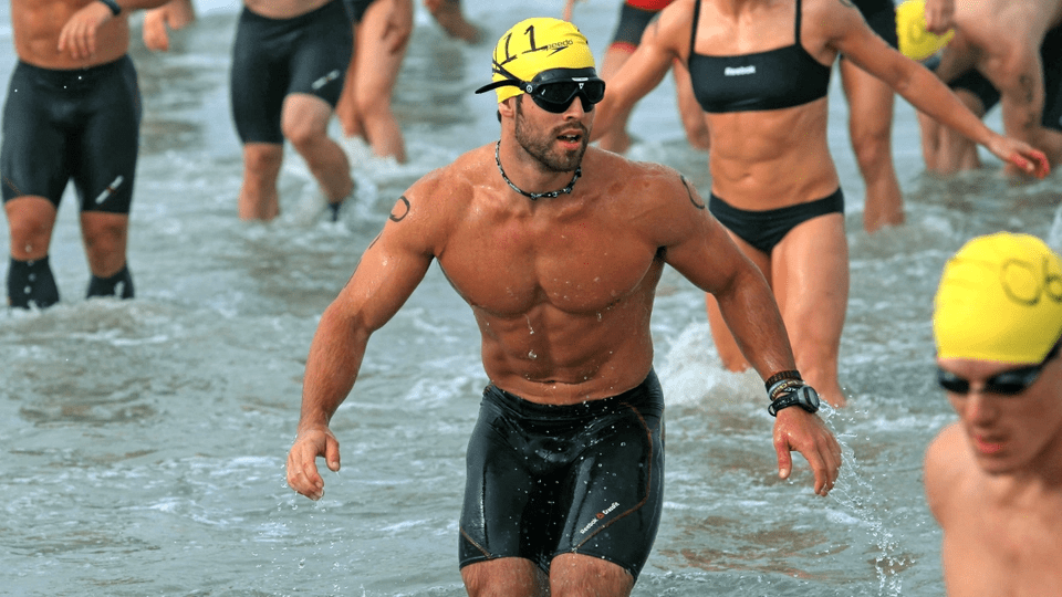 Male swimmer in a yellow cap exits the ocean, surrounded by competitors. The scene captures the intense atmosphere of a triathlon event.