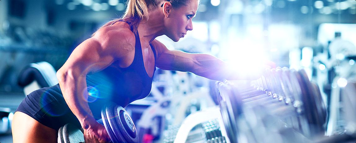 A focused woman lifts weights in a gym, showcasing strength and determination. The bright lighting highlights her muscular form and dedication.